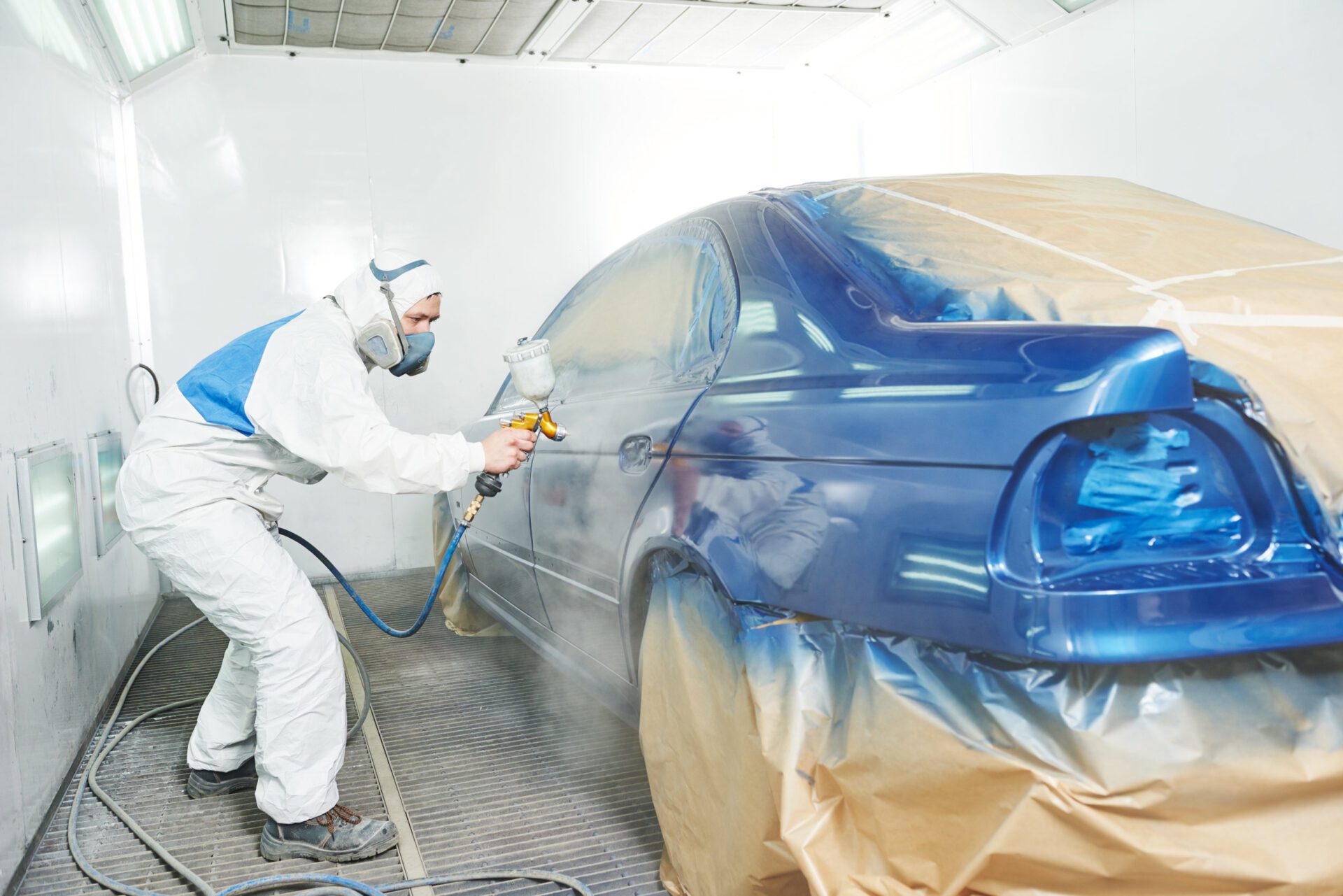 worker painting auto car body automobile repairman painter in protective workwear and respirator painting car body bumper in paint chamber